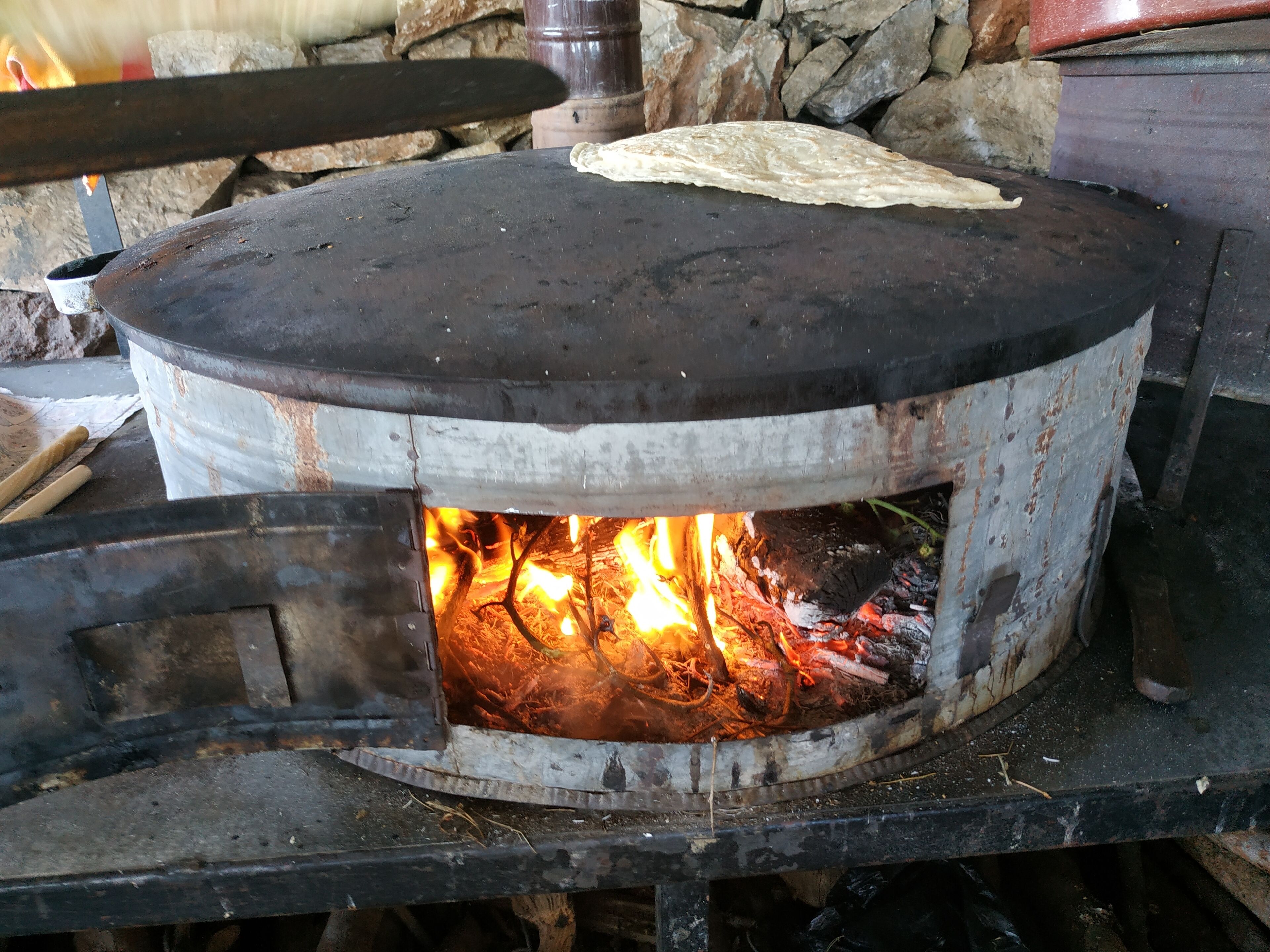 A traditional wood-fired oven with a flatbread on top, surrounded by stone walls and a rustic setting.