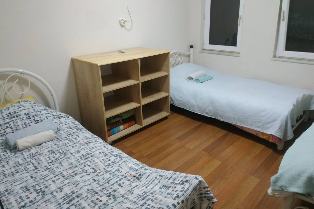 A small, tidy room with two single beds, a wooden bookshelf, and a window.