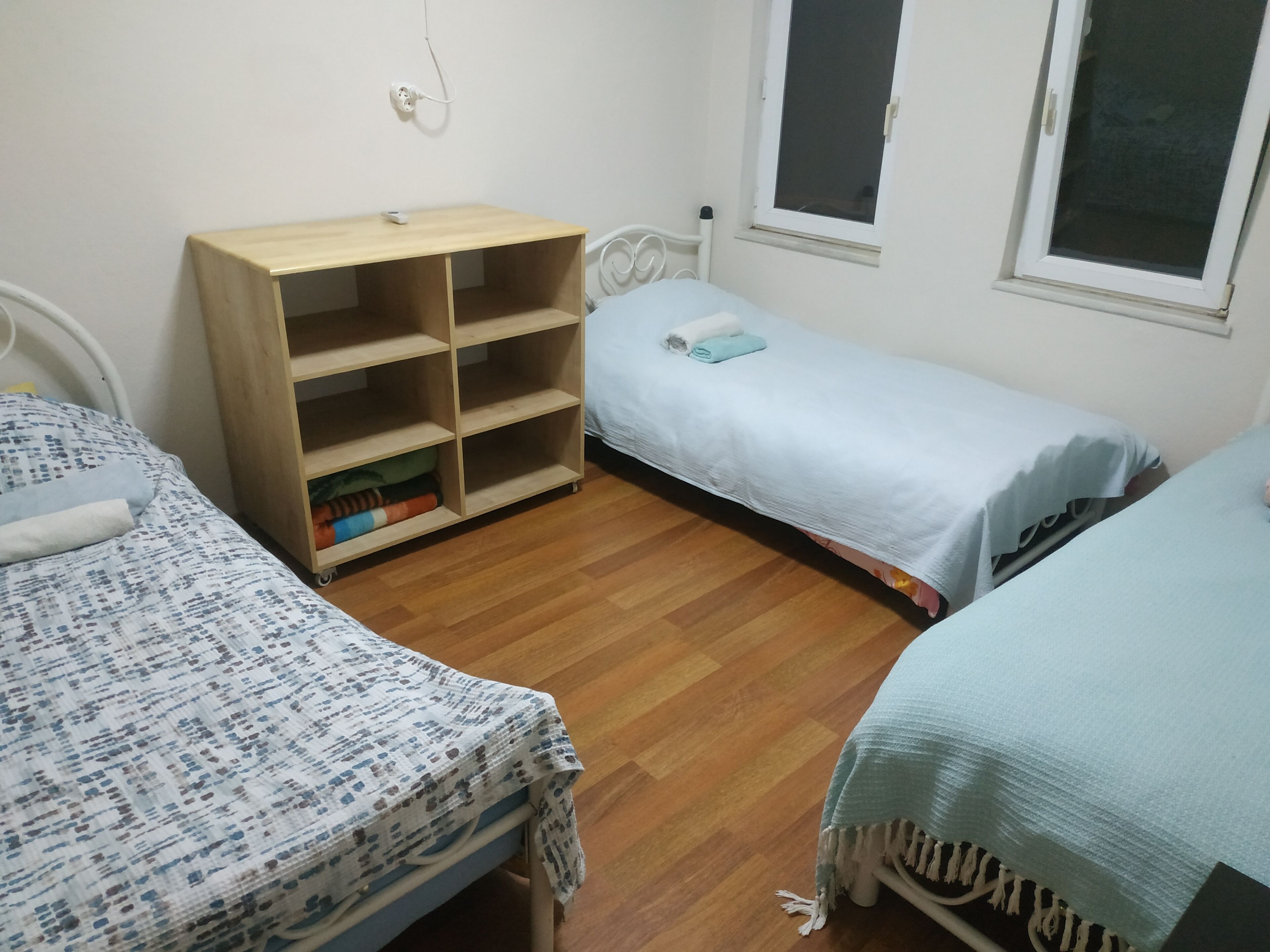 Une petite pièce propre et bien rangée, avec trois lits, une étagère en bois et une fenêtre.
