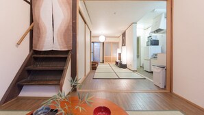 A traditional Japanese interior with tatami flooring, a low wooden table with a red bowl, a staircase with a wooden handrail, and a kitchen in the background. - Kyo Machiya Nijo (Kyoto)