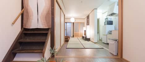 Un interior japonés tradicional con suelo de tatami, una mesa de madera baja con un tazón rojo, una escalera con barandilla de madera y una cocina al fondo.