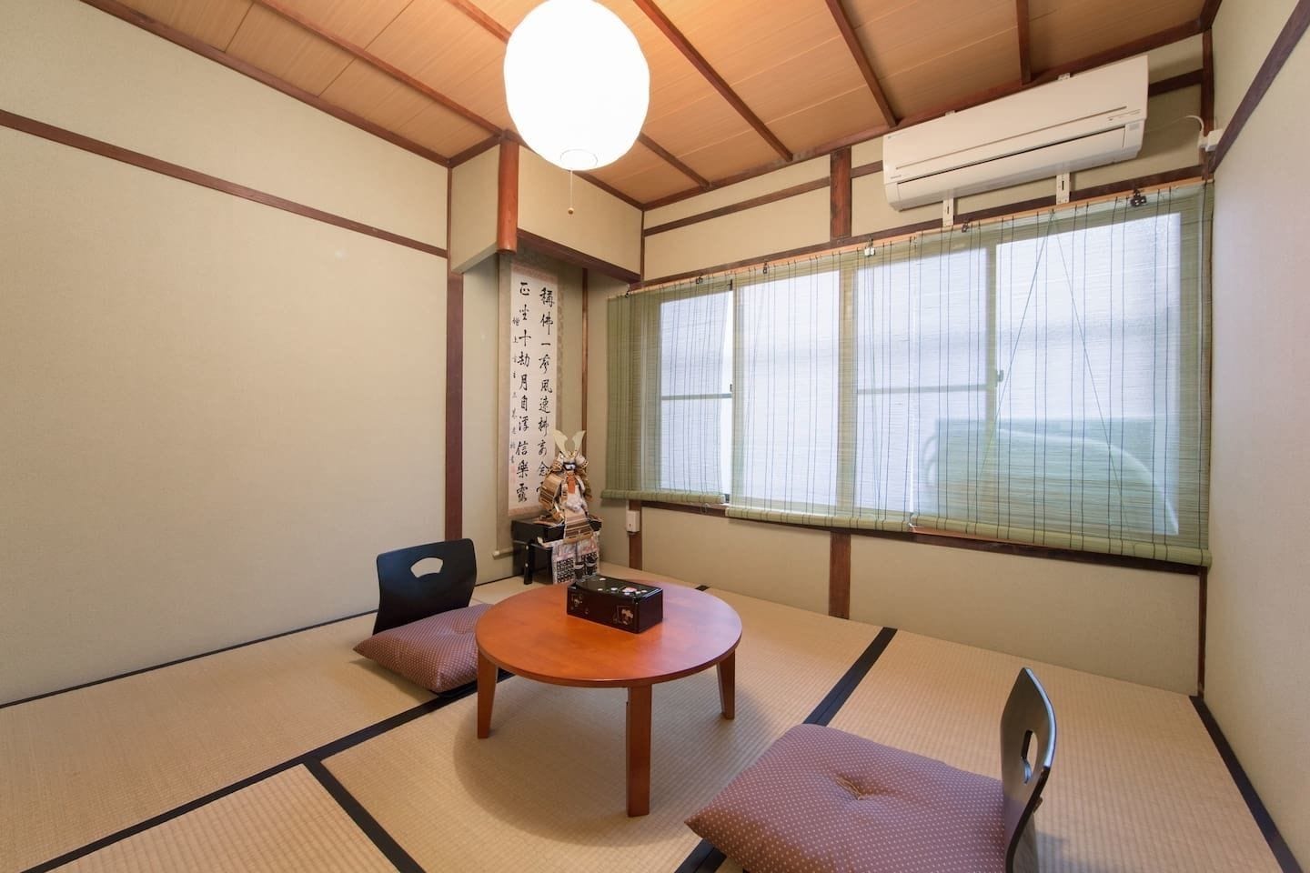 A traditional Japanese room with tatami flooring, a low table, a cushion, a decorative figure, and a window with blinds.
