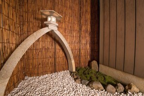 Une lanterne en pierre surmontée d’une arche incurvée, un mur en bambou et un sol recouvert de petits cailloux, de mousse et de rochers.