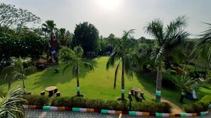 A lush garden with palm trees, a grassy lawn, and a paved walkway. - OVER NIGHT STAY ARAVALI ADVENTURE, GURGAON DELHI NCR (Gurugram)