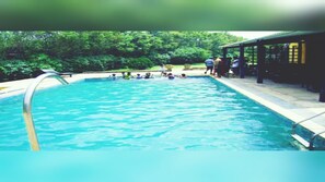 A swimming pool with clear blue water, surrounded by a tiled deck and a covered area with people. - OVER NIGHT STAY ARAVALI ADVENTURE, GURGAON DELHI NCR (Gurugram)
