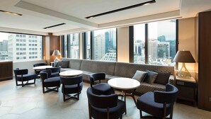 A modern lounge area with a sofa, chairs, and tables, large windows with a city view, and a lamp on a side table. - Sotetsu Fresa Inn Seoul Myeong Dong (Seoul)