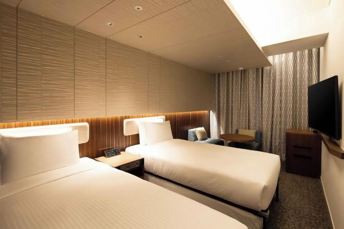 Habitación de hotel con dos camas, televisión, una mesita y ventana con cortinas.