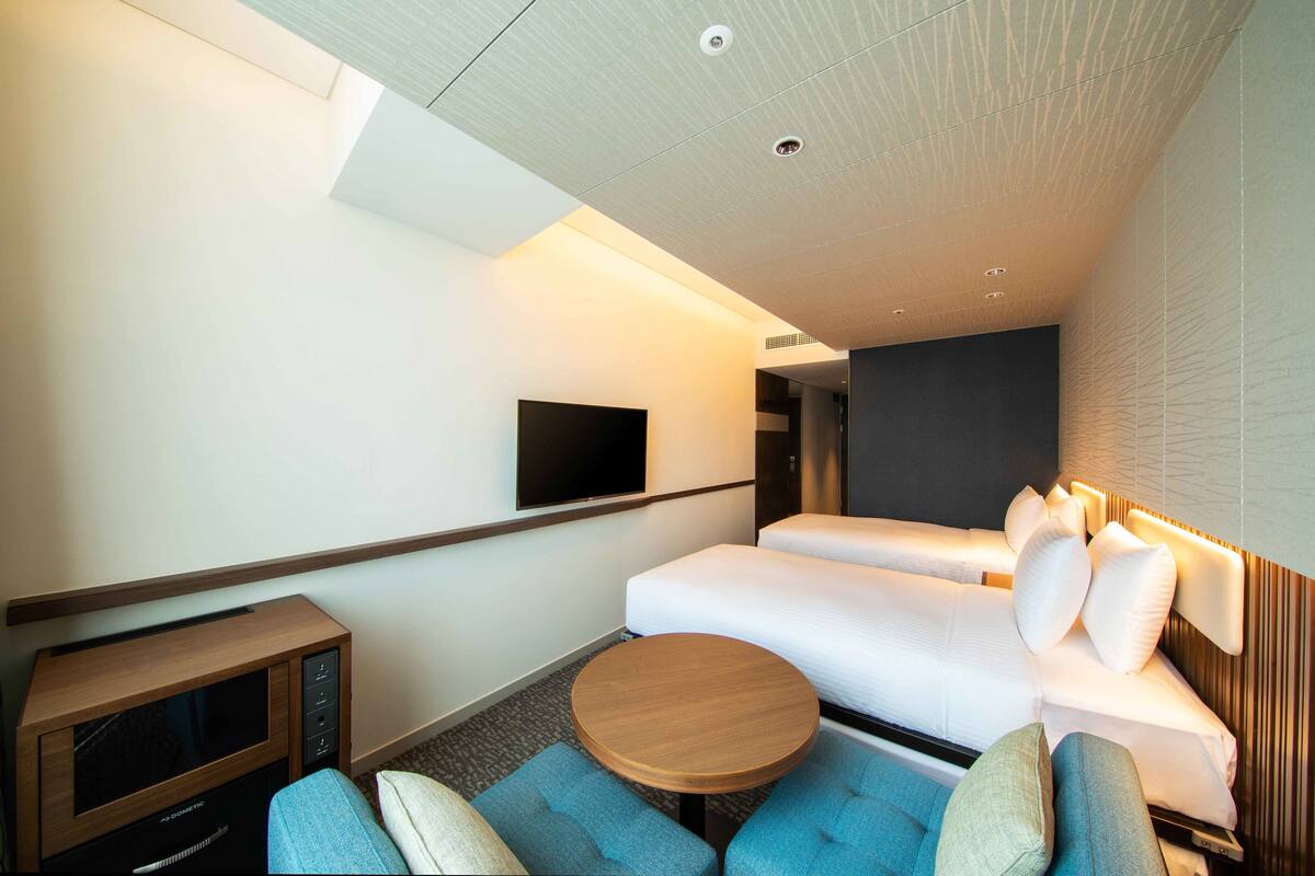 Une chambre d’hôtel moderne équipée d’un téléviseur à écran plat fixé au mur, d’un canapé-lit avec des coussins, d’un petit meuble en bois pour la télévision et d’une table ronde en bois.