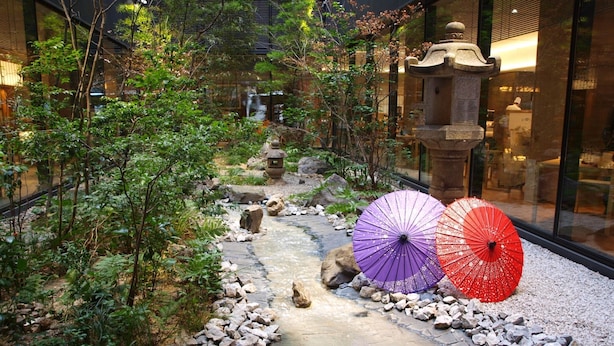 A pathway lined with rocks and greenery, leading to a traditional stone lantern and a building with large windows.