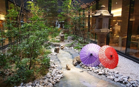 A pathway lined with rocks and greenery, leading to a traditional stone lantern and a building with large windows.