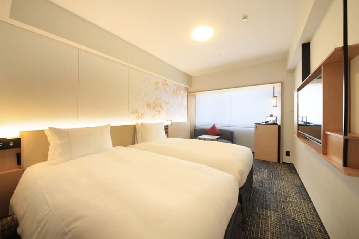 A hotel room with two beds, a small desk, a chair, a window with a view, and a wall-mounted TV.