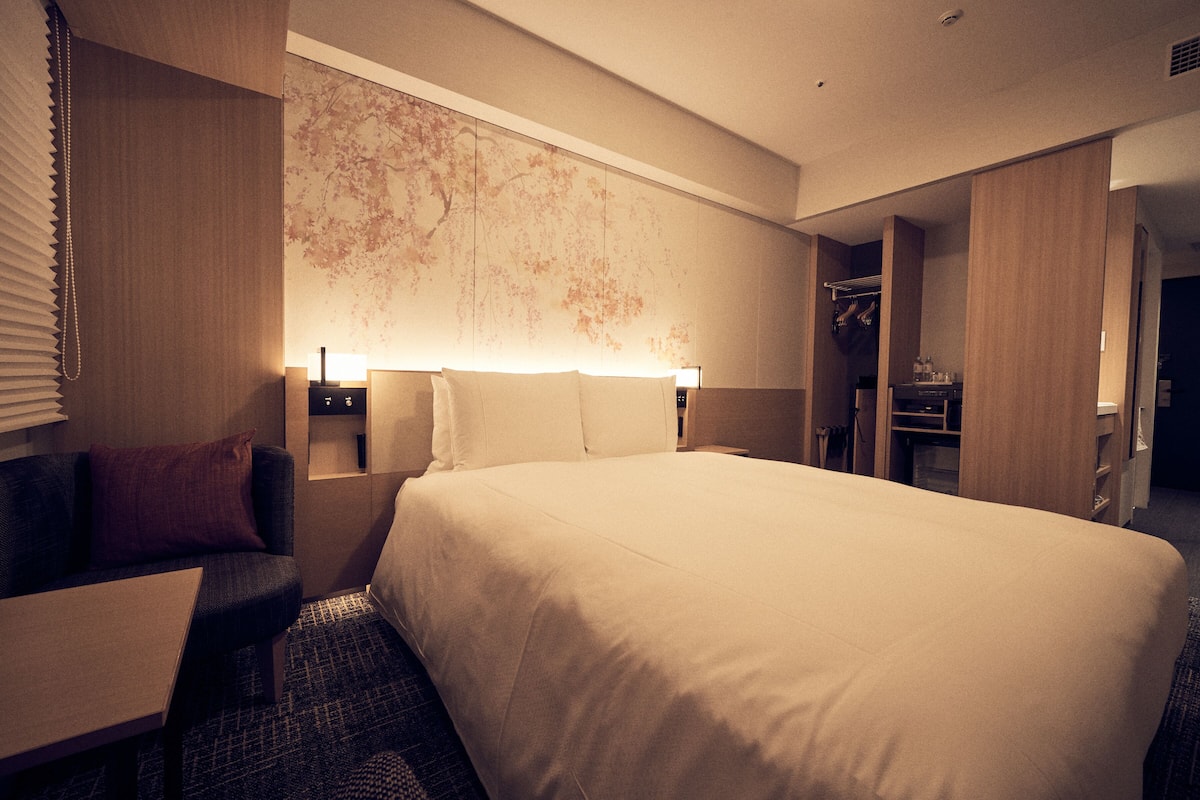 A hotel room with a large bed, a desk, a chair, a wardrobe, and a wall-mounted lamp.