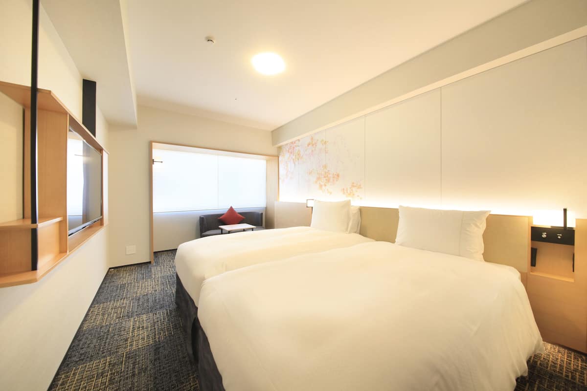 A hotel room with two beds, a small table, a chair, and a window with a view.