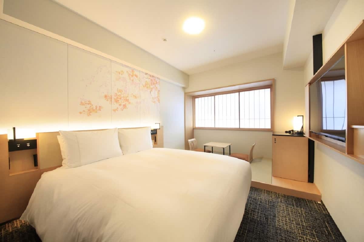 A hotel room with a large bed, a small table, a chair, a desk, and a large window.