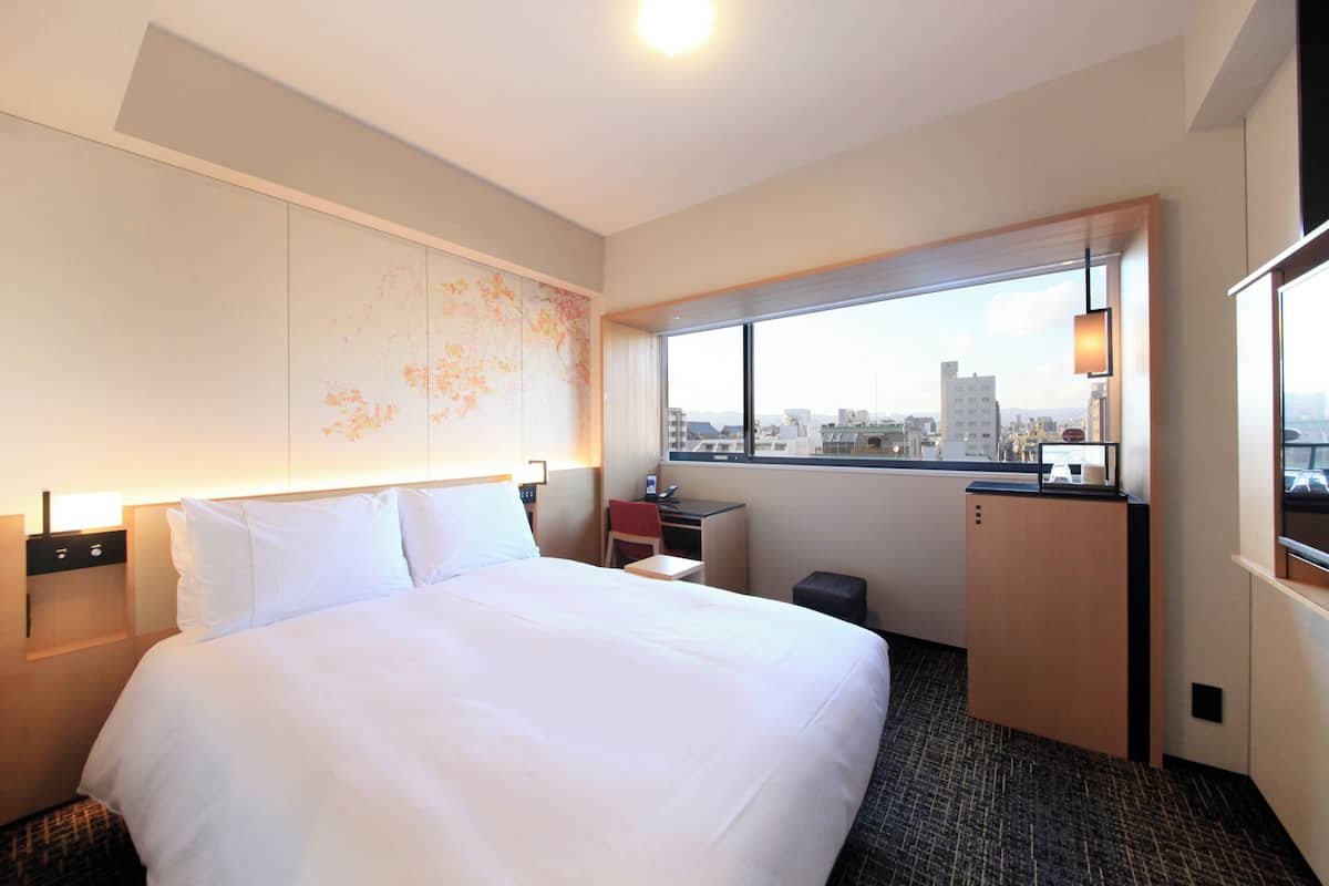 A hotel room with a large bed, a desk, a chair, a small table, a window with a city view, and a minibar.