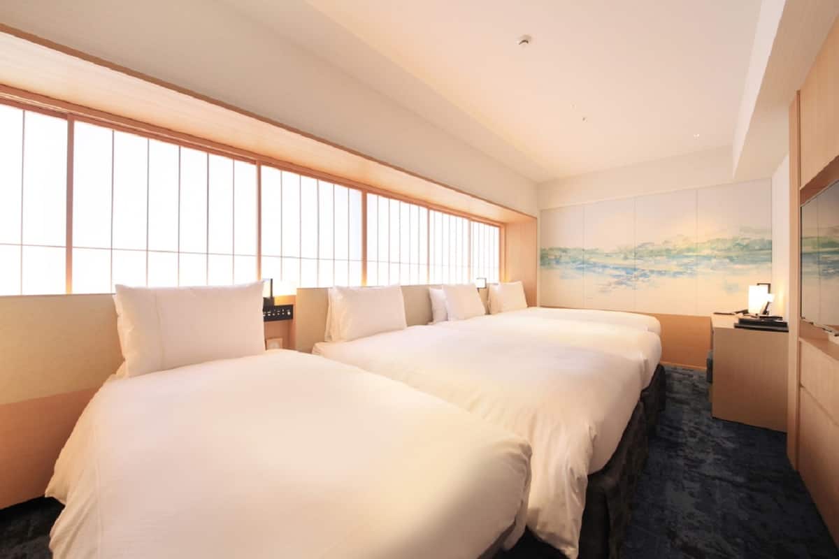 A hotel room with two beds, a large window with blinds, a painting on the wall, and a desk with a lamp.