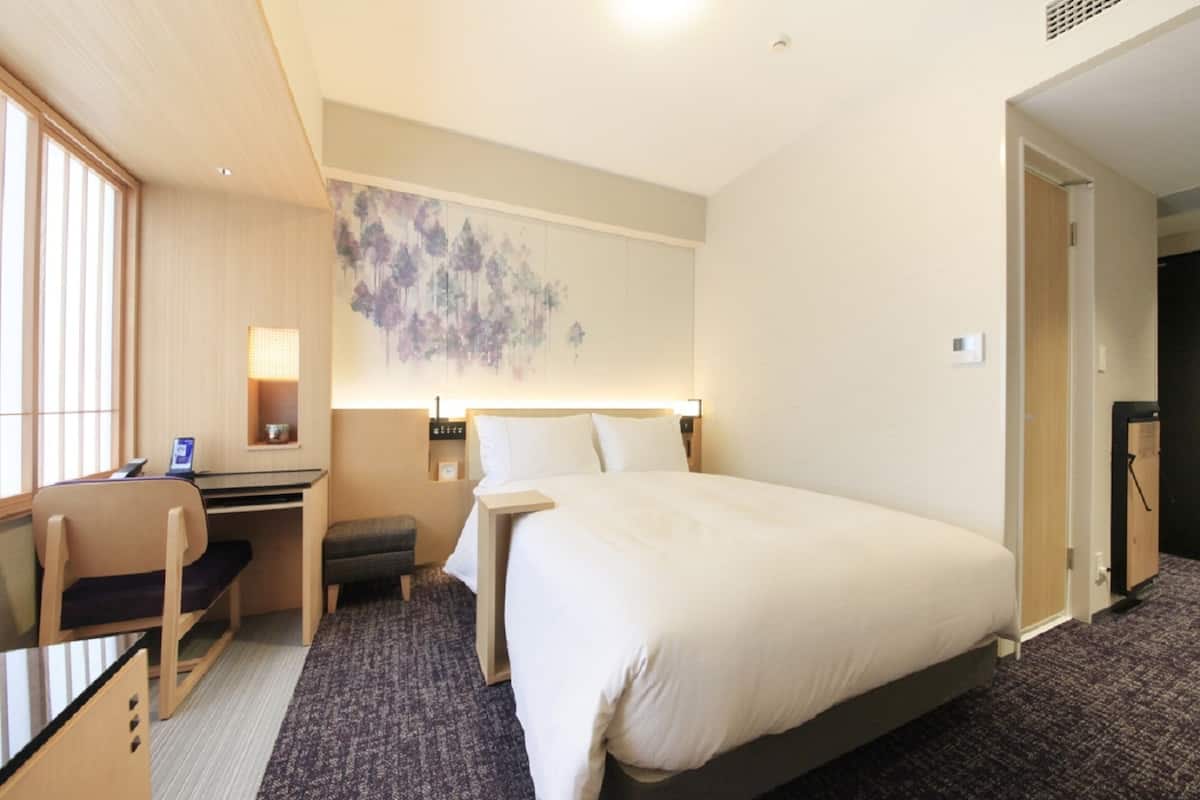 A hotel room with a bed, desk, chair, and a wall-mounted artwork.