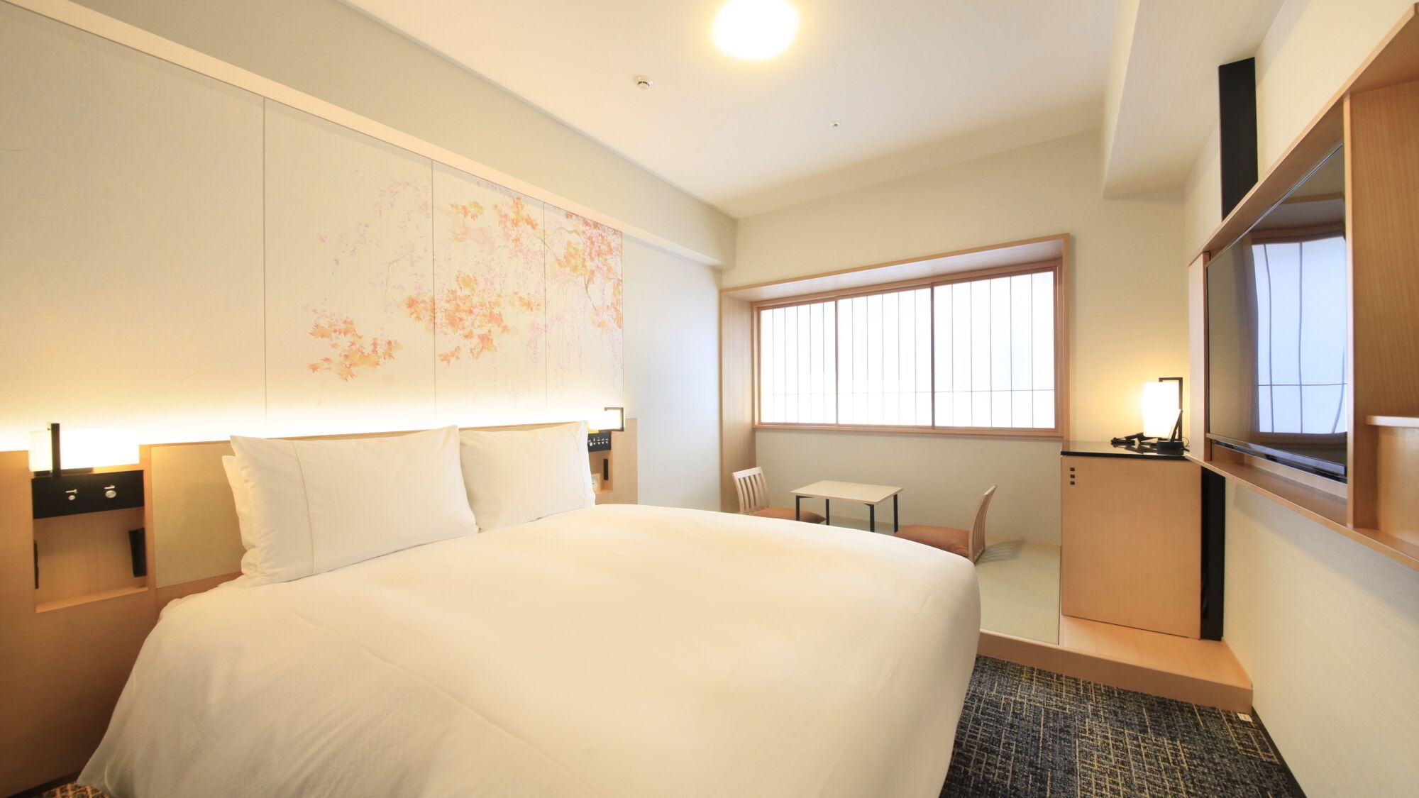 A hotel room with a large bed, a small table, a chair, a desk, a TV, and a window.