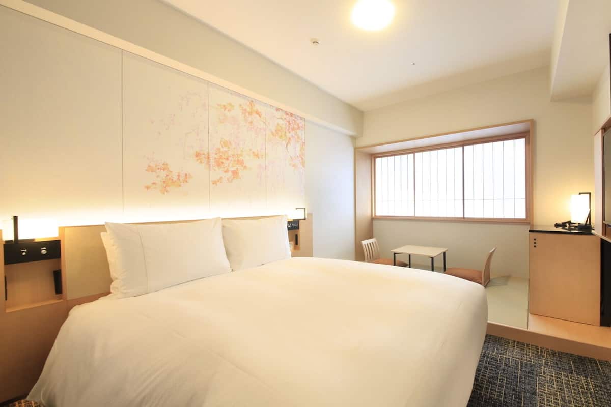 A hotel room with a large bed, a small table, a chair, a desk, a TV, and a window.