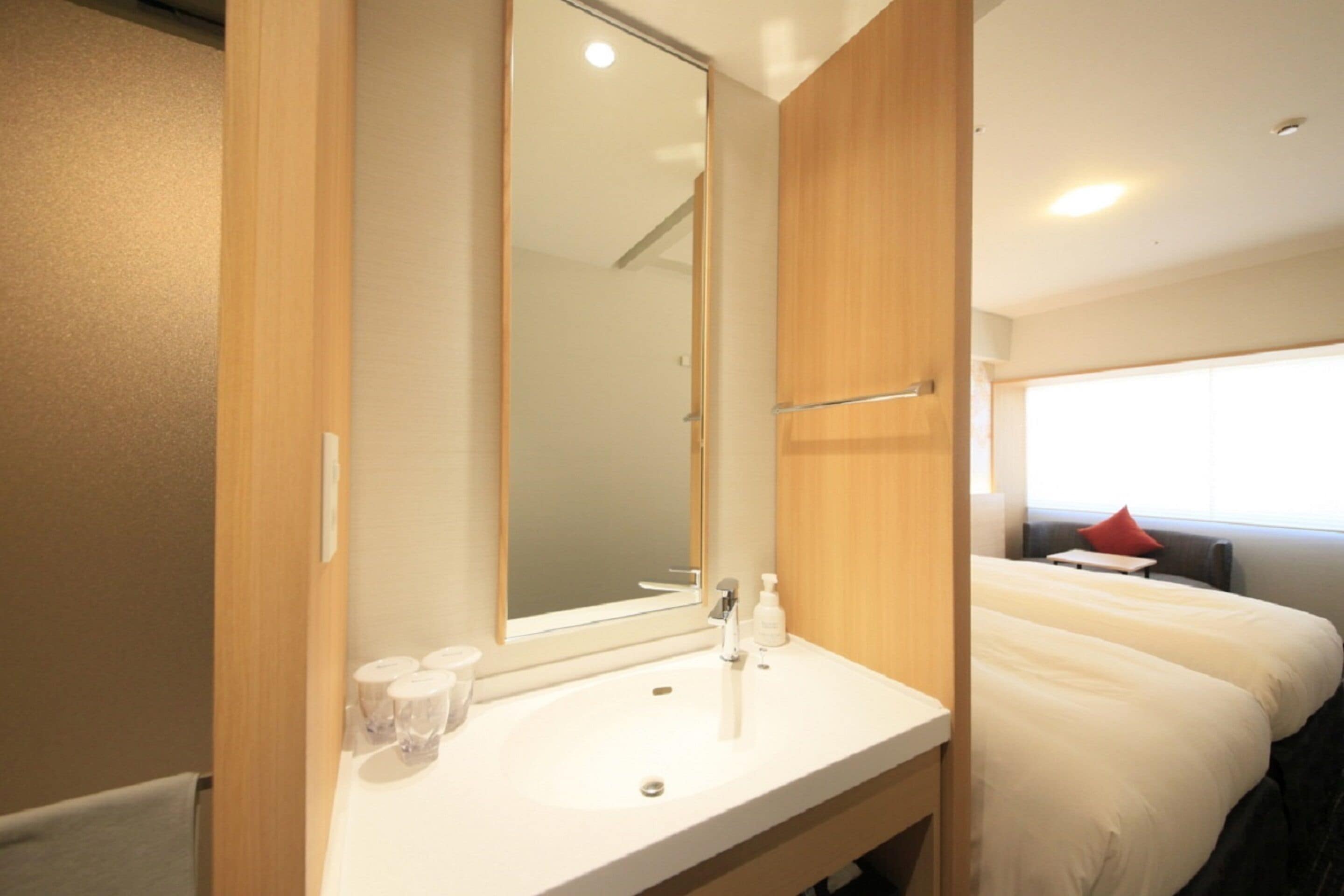 A hotel room with a large mirror, a white sink, a bed with white bedding, and a window with a red pillow.