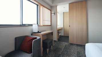 A hotel room with a bed, a desk, a chair, a small stool, a window with blinds, and a wardrobe.