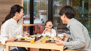 A dining table with various dishes and drinks, a window with a view of the street, and a person holding chopsticks.