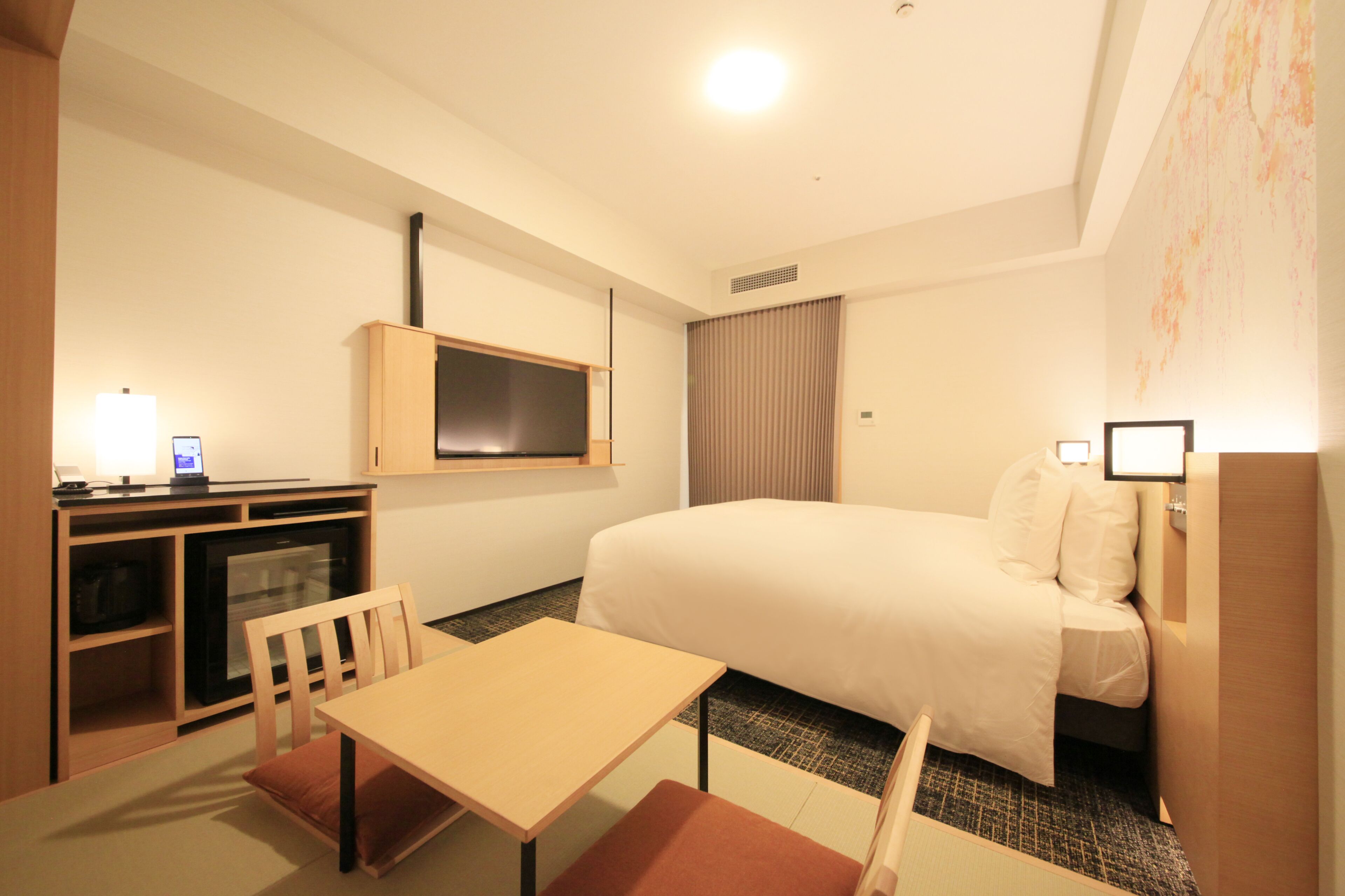 A hotel room with a bed, a desk, a chair, a television, and a small kitchenette.