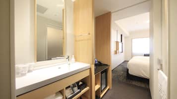 A hotel room with a large mirror, a white sink, a bedside table with a phone, a bed, and a window.
