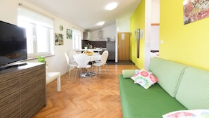 Ein modernes Wohnzimmer mit Flachbildfernseher, einem grünen Sofa, einem Essbereich mit weißem Tisch und Stühlen sowie einer Küche im Hintergrund.