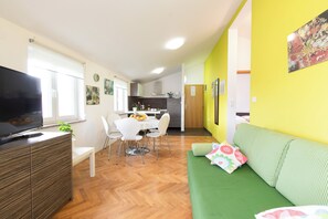 A modern living room with a flat-screen TV, a green sofa, a dining area with a white table and chairs, and a kitchen in the background.