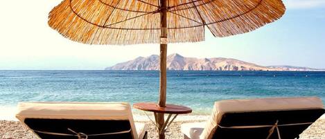 A beach scene with two lounge chairs under a thatched umbrella, a small round table, and a view of the sea and distant mountains.