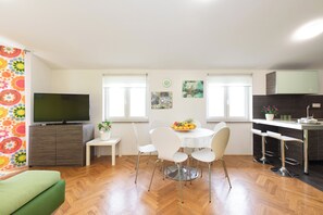 A modern living room with a flat-screen TV, a dining table with chairs, a sofa, and a kitchen area with a bar stool.