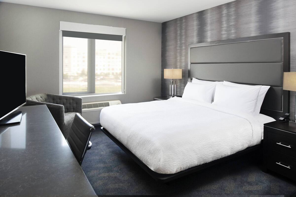 A modern hotel room with a large bed, a desk, and a chair. There is a window providing a view of buildings outside.