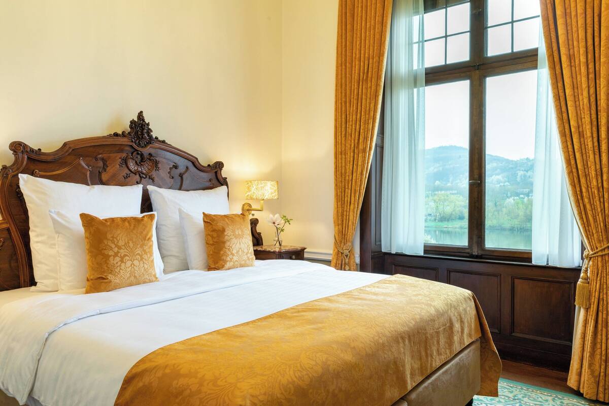 A large bed with a wooden headboard, white and patterned pillows, and a gold bedspread. There is a nightstand with a lamp and a vase of flowers. A large window with curtains provides a view of a landscape.