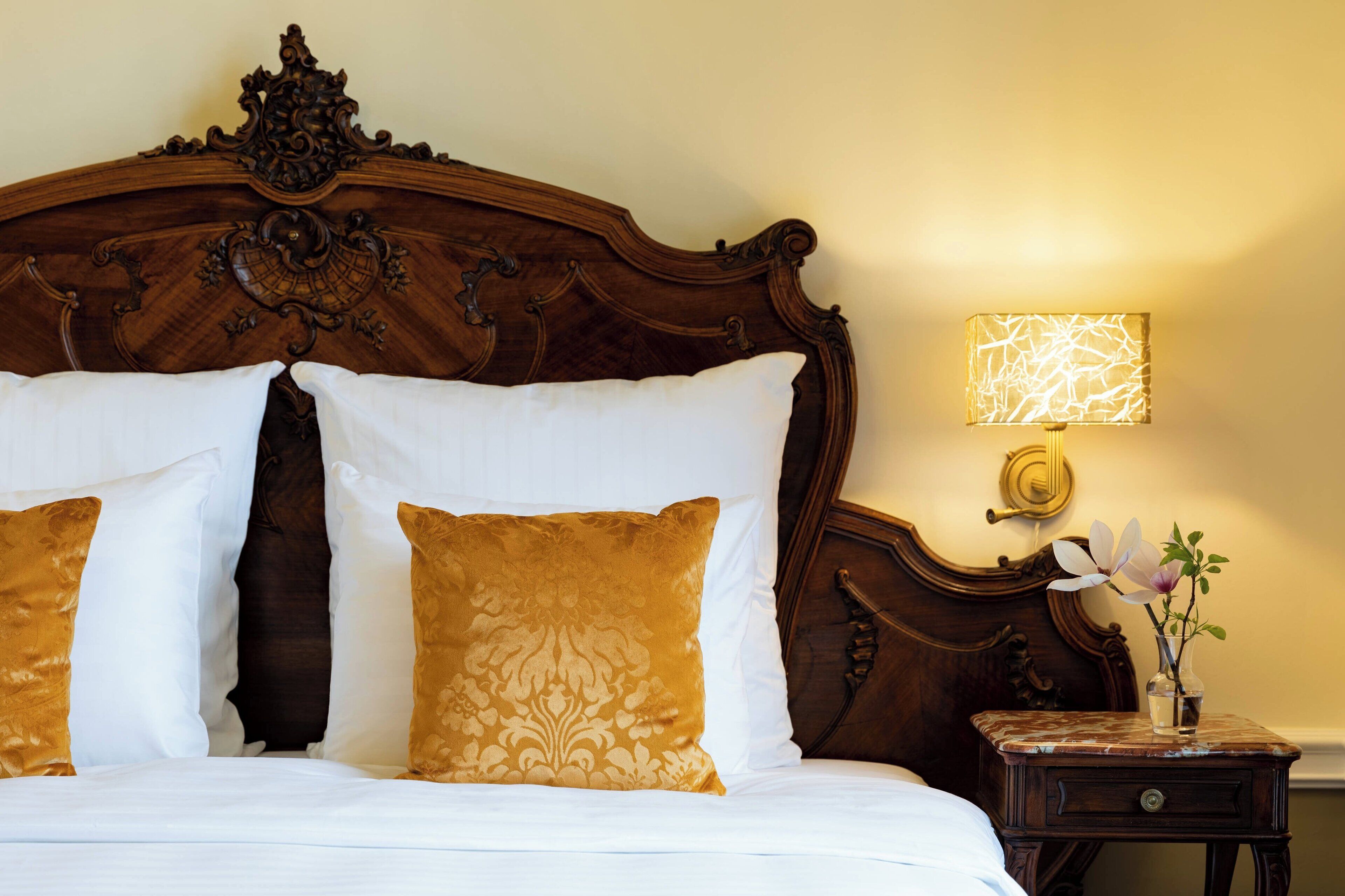 Ein Bett mit weißen und goldenen Kissen, einem Holz-Kopfteil und einem Nachttisch mit Lampe und Blumenvase.