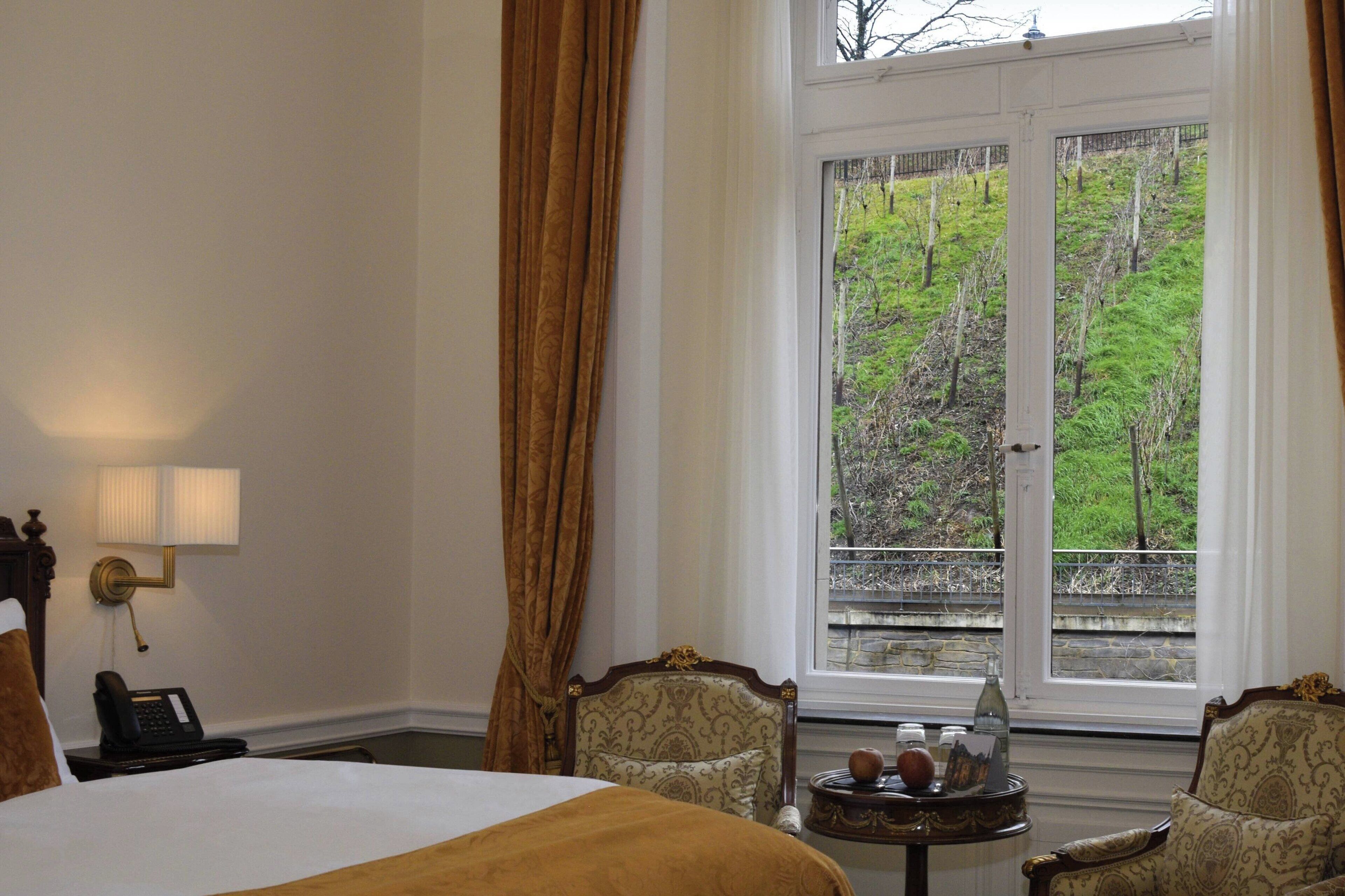 Ein Hotelzimmer mit einem großen Bett, zwei Sesseln, einem kleinen Tisch mit einer Vase und einem Fenster mit Blick auf eine grüne Landschaft.
