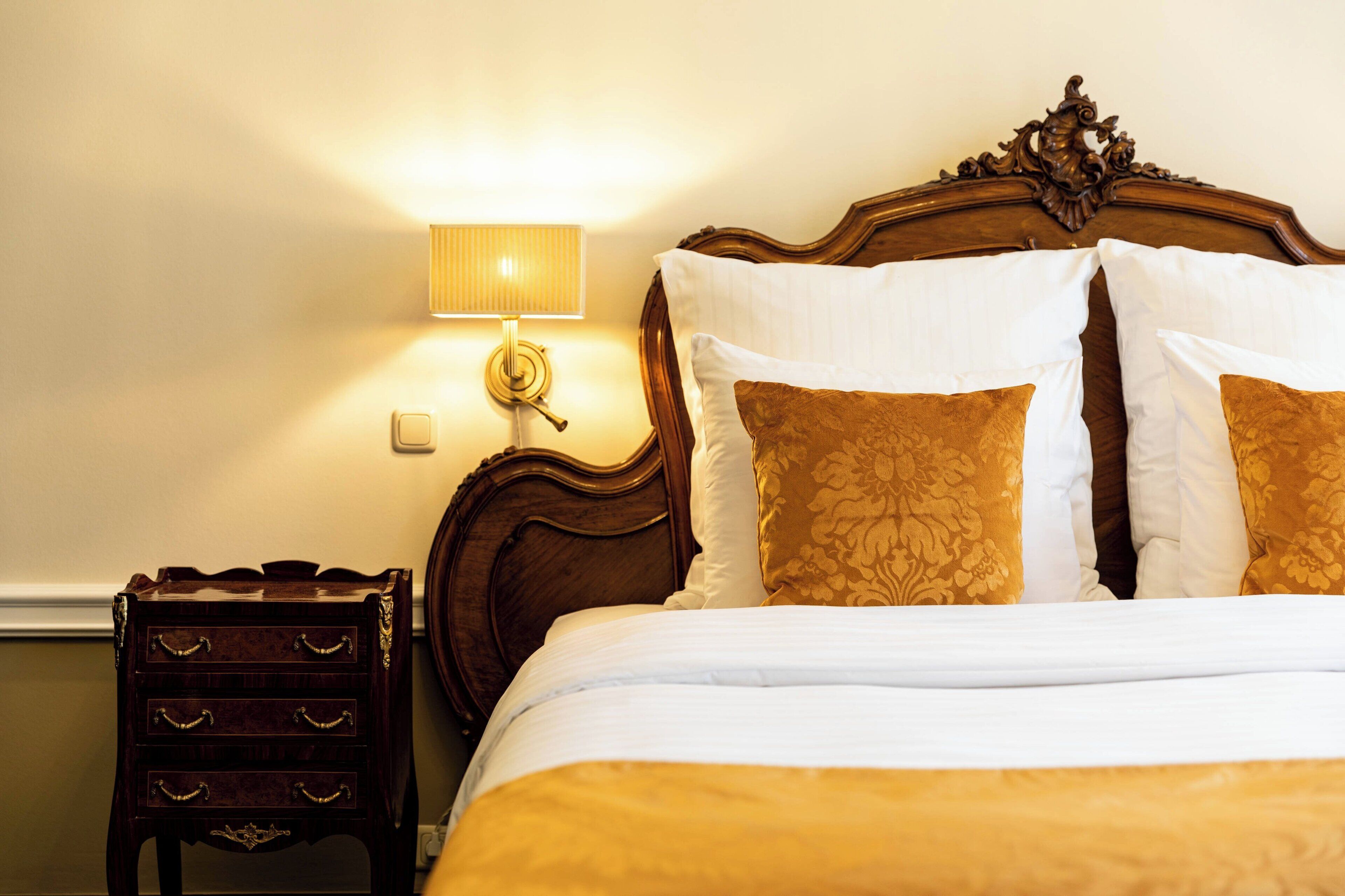 A neatly made bed with white and gold bedding, a wooden headboard, and a bedside table with drawers.