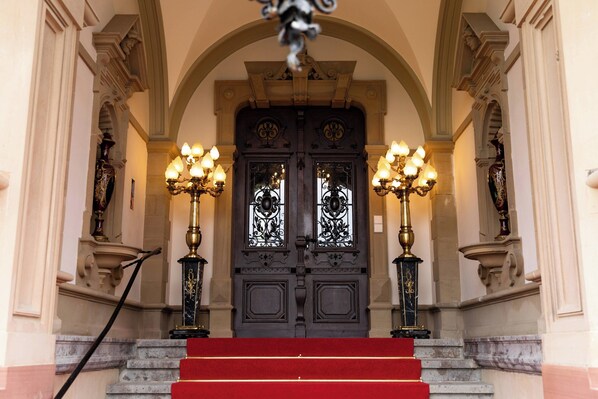 Una entrada imponente con alfombra roja que conduce a una puerta de madera oscura, flanqueada por candelabros negros y dorados de gran detalle.