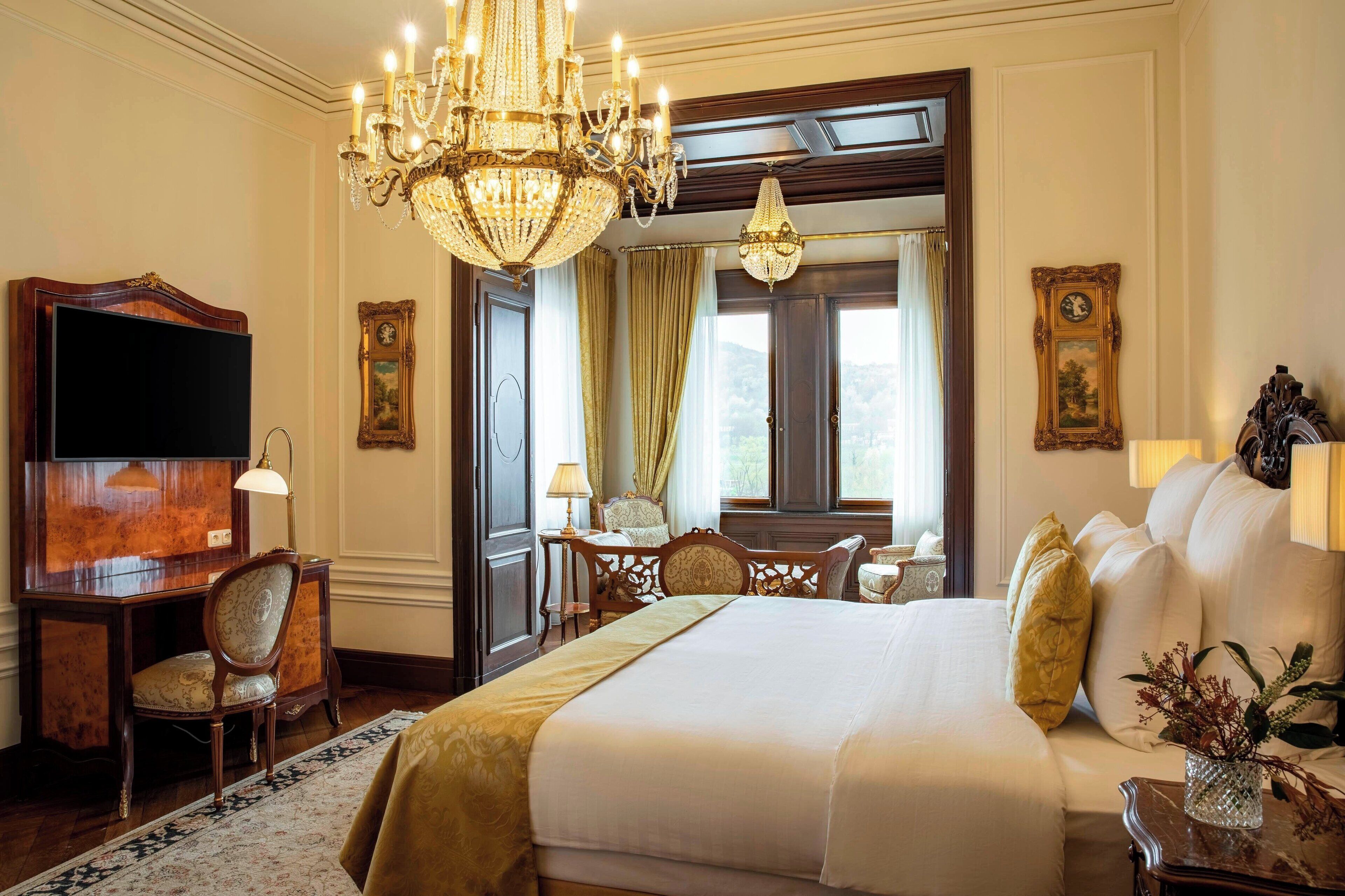 A hotel room with a large bed, a desk with a television, a chandelier, and a view of the outdoors.