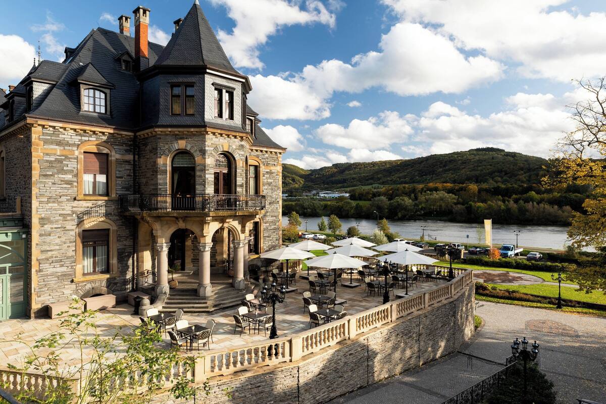 A large stone building with a balcony overlooking a river, surrounded by outdoor seating and umbrellas.