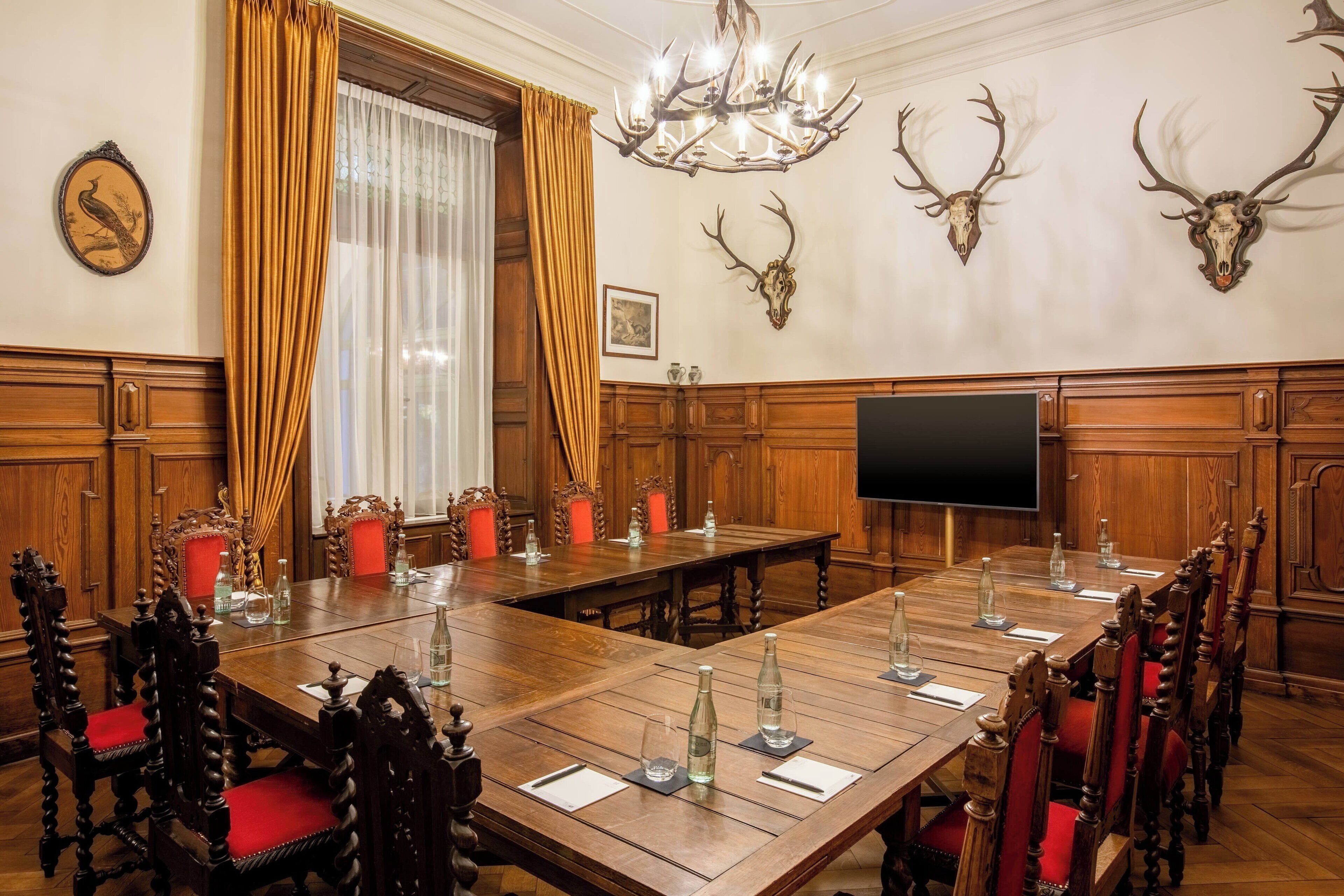 A conference room with a large wooden table, red upholstered chairs, a chandelier, and mounted antler heads on the wall.