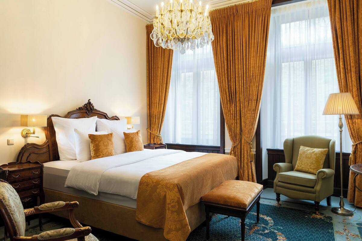 A hotel room with a large bed, a sitting area with a chair and a table, a chandelier, and curtains.
