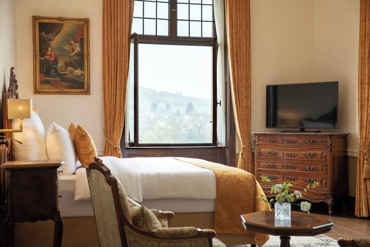 A hotel room with a large bed, a television, a wooden dresser, and a window with a view of a landscape.