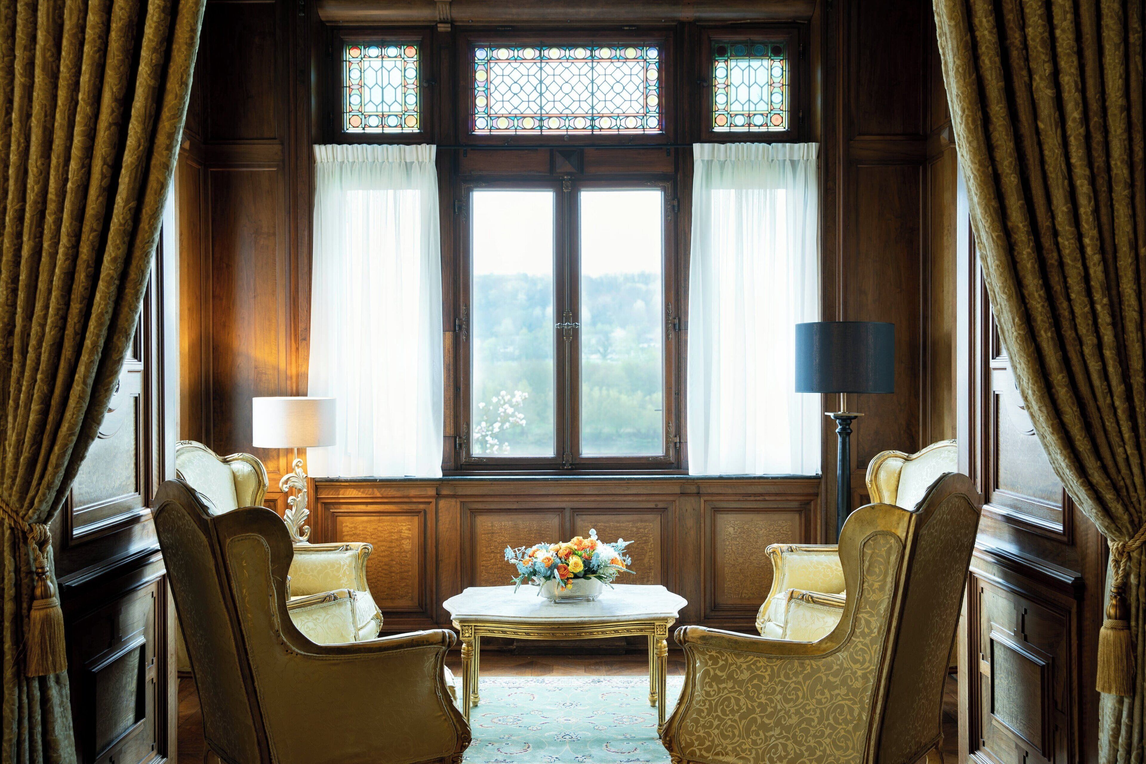 A room with two chairs, a table with a floral arrangement, and a view through a large window with curtains.