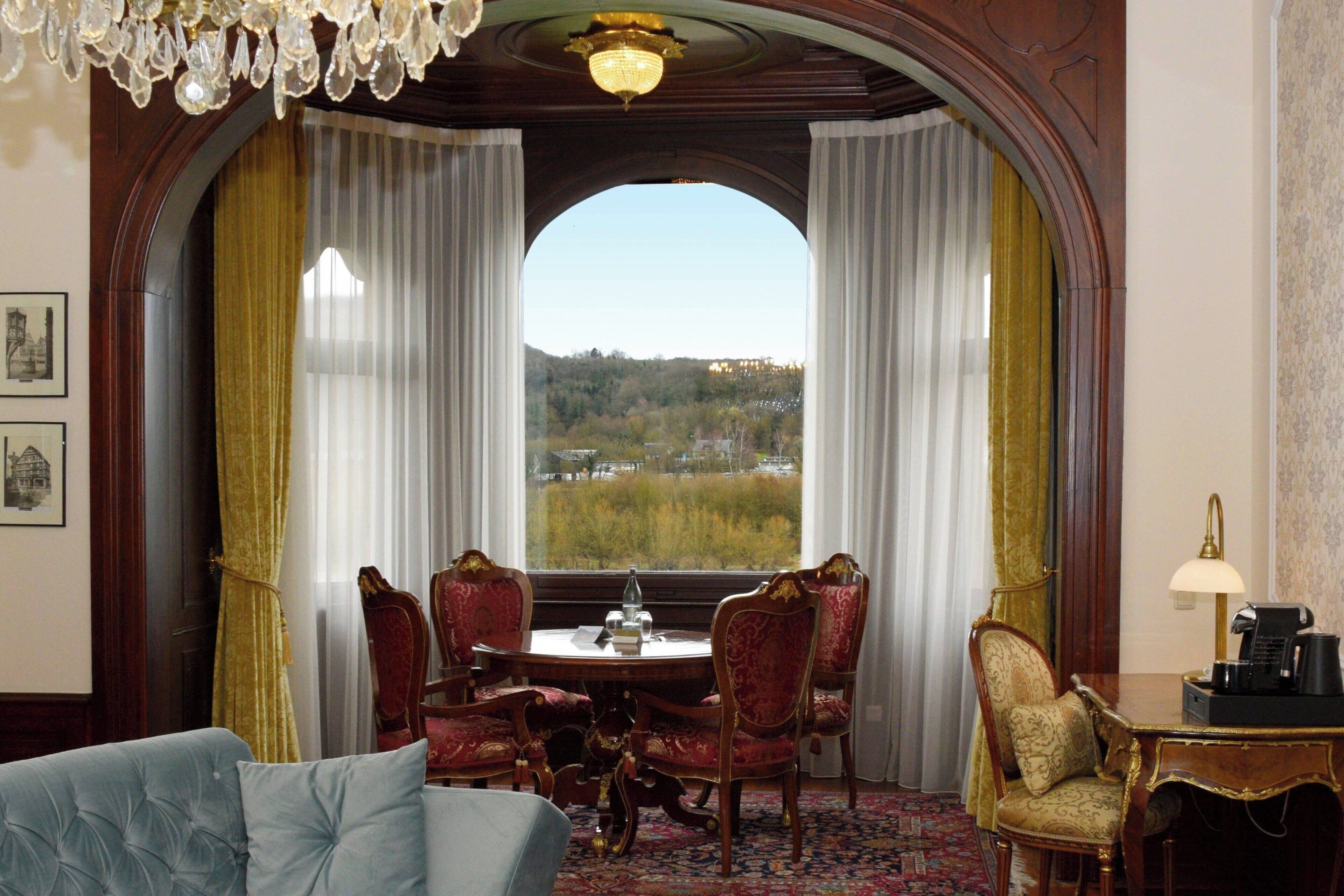 A room with a dining table and chairs, a chandelier, and a view of a landscape through a large window.