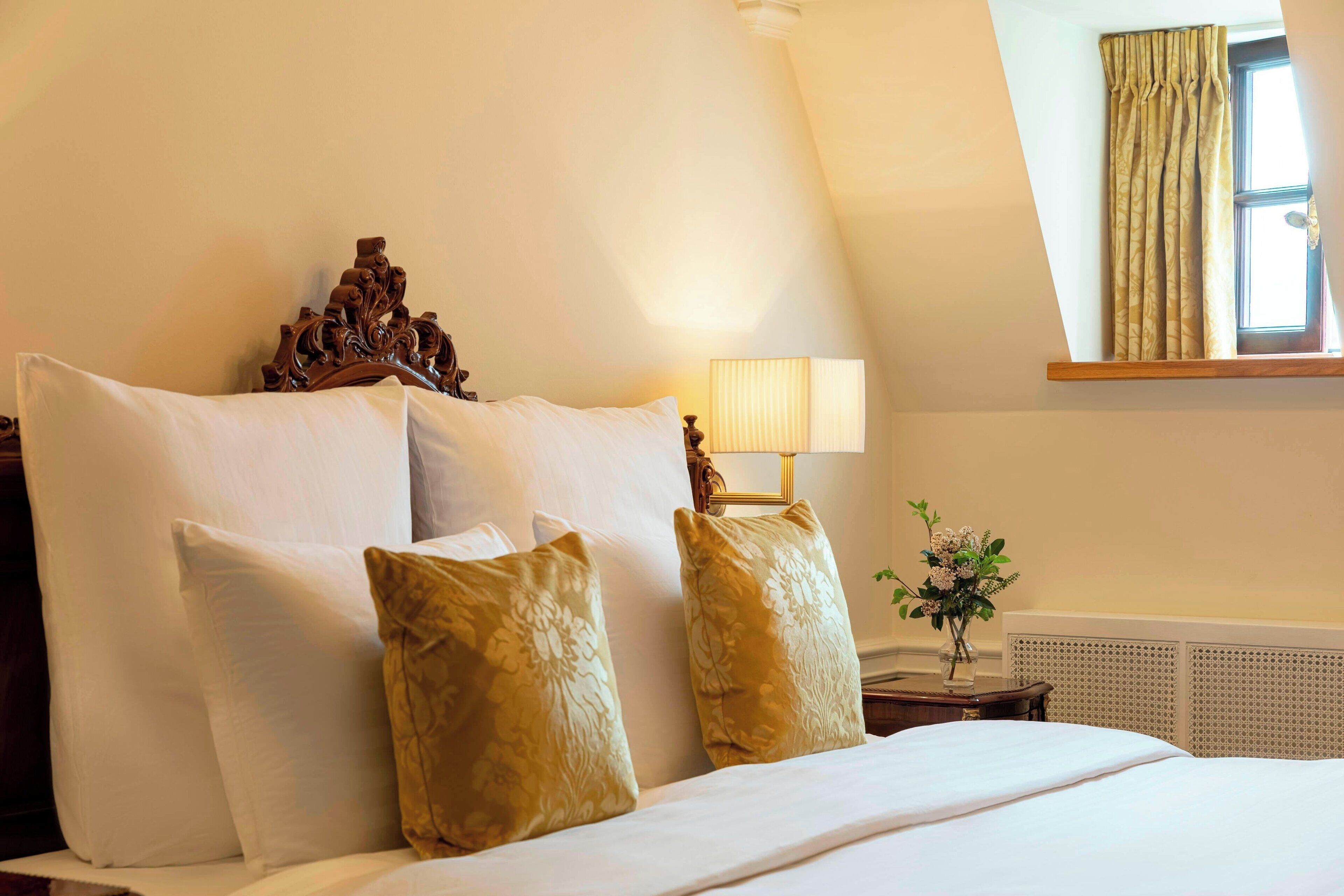 A neatly made bed with white and gold pillows, a wooden headboard, and a bedside lamp.
