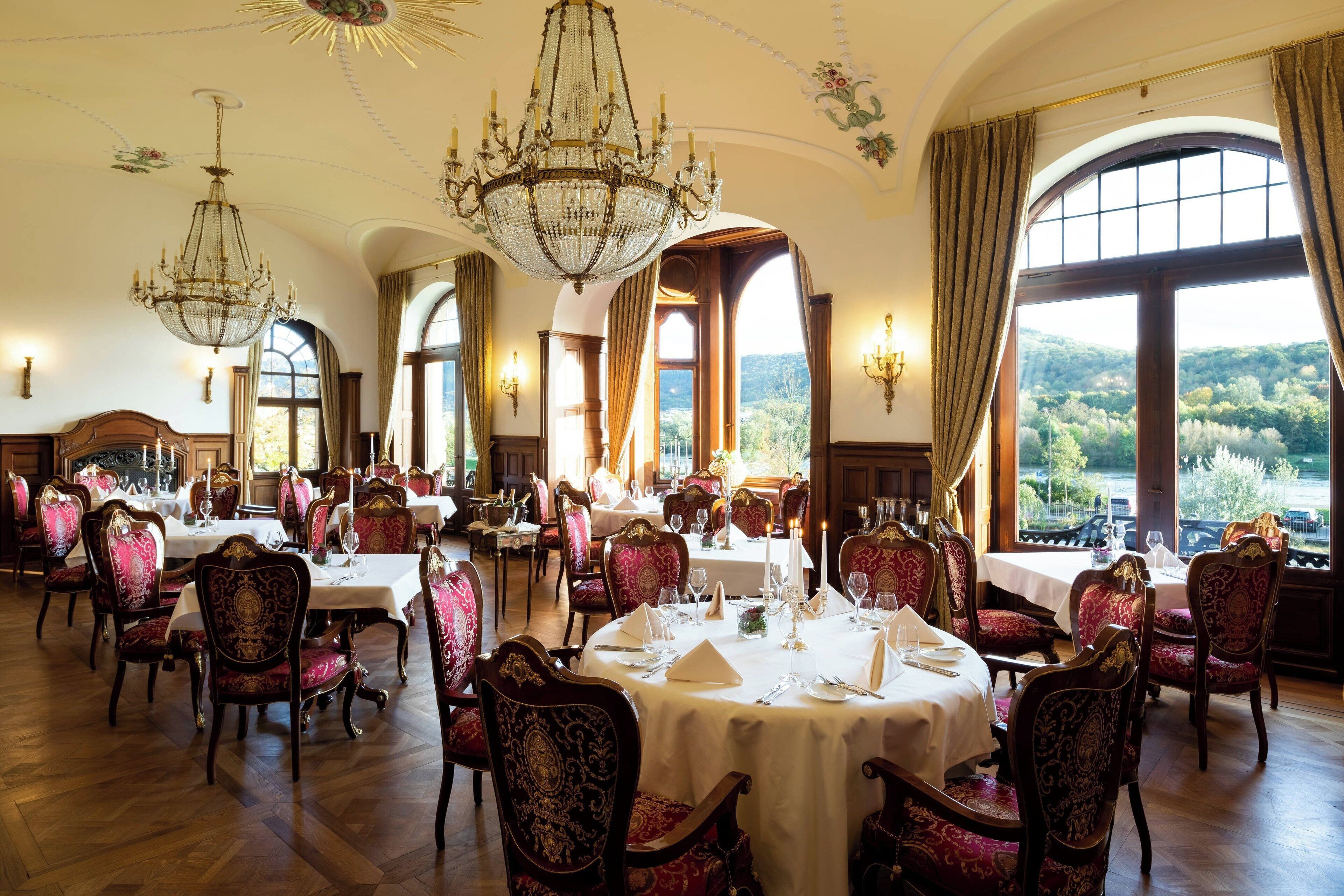 Un comedor amplio y elegante con candelabros ornamentados, sillas tapizadas en rojo y mesas dispuestas para comer.