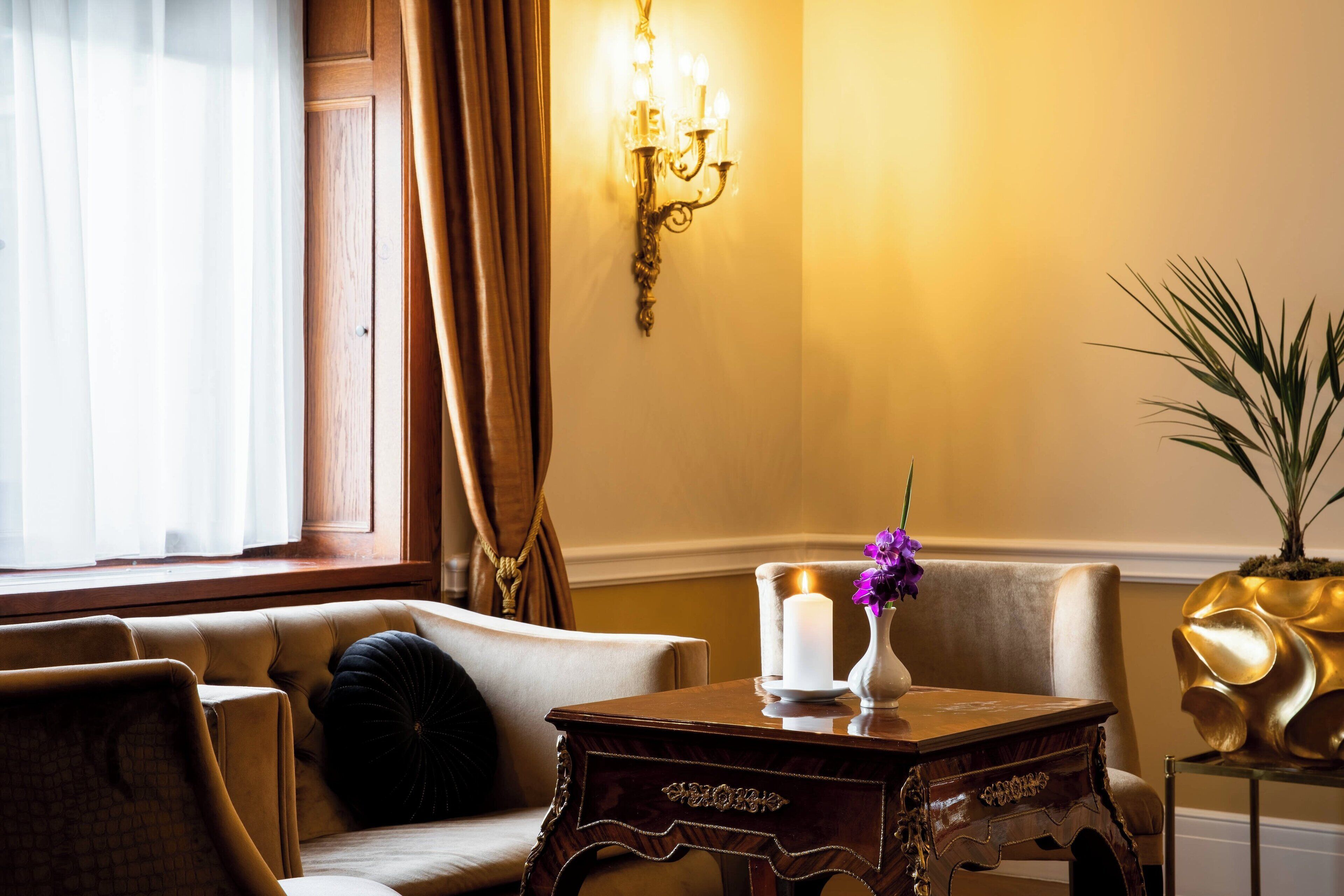 A room with a sofa, a table with a candle and flowers, a wall-mounted light fixture, and a potted plant.