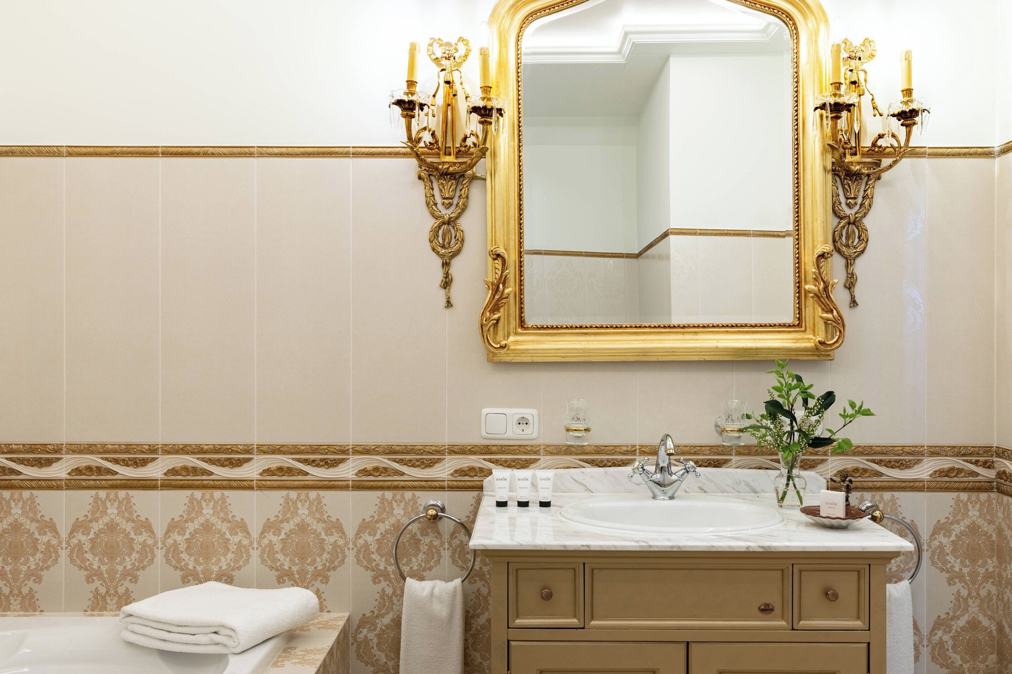 A bathroom with a large mirror, gold sconces, a sink with toiletries, and a towel ring.