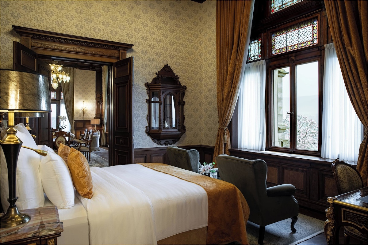 Una habitación con una cama grande, una silla de madera, una mesa con una lámpara y un ventanal con cortinas.